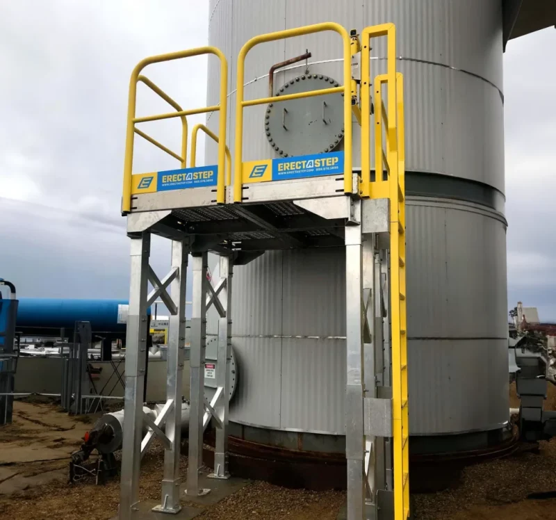 Custom ladder platform installed to provide safe access to a tank access door in a facility, featuring a compact design and secure footing.